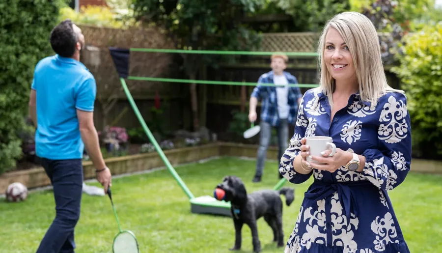Laura staat in haar tuin terwijl haar familie op de achtergrond badminton speelt. Met Peristeen Plus houdt ze haar darmklachten, die door MS veroorzaakt worden, onder controle. 